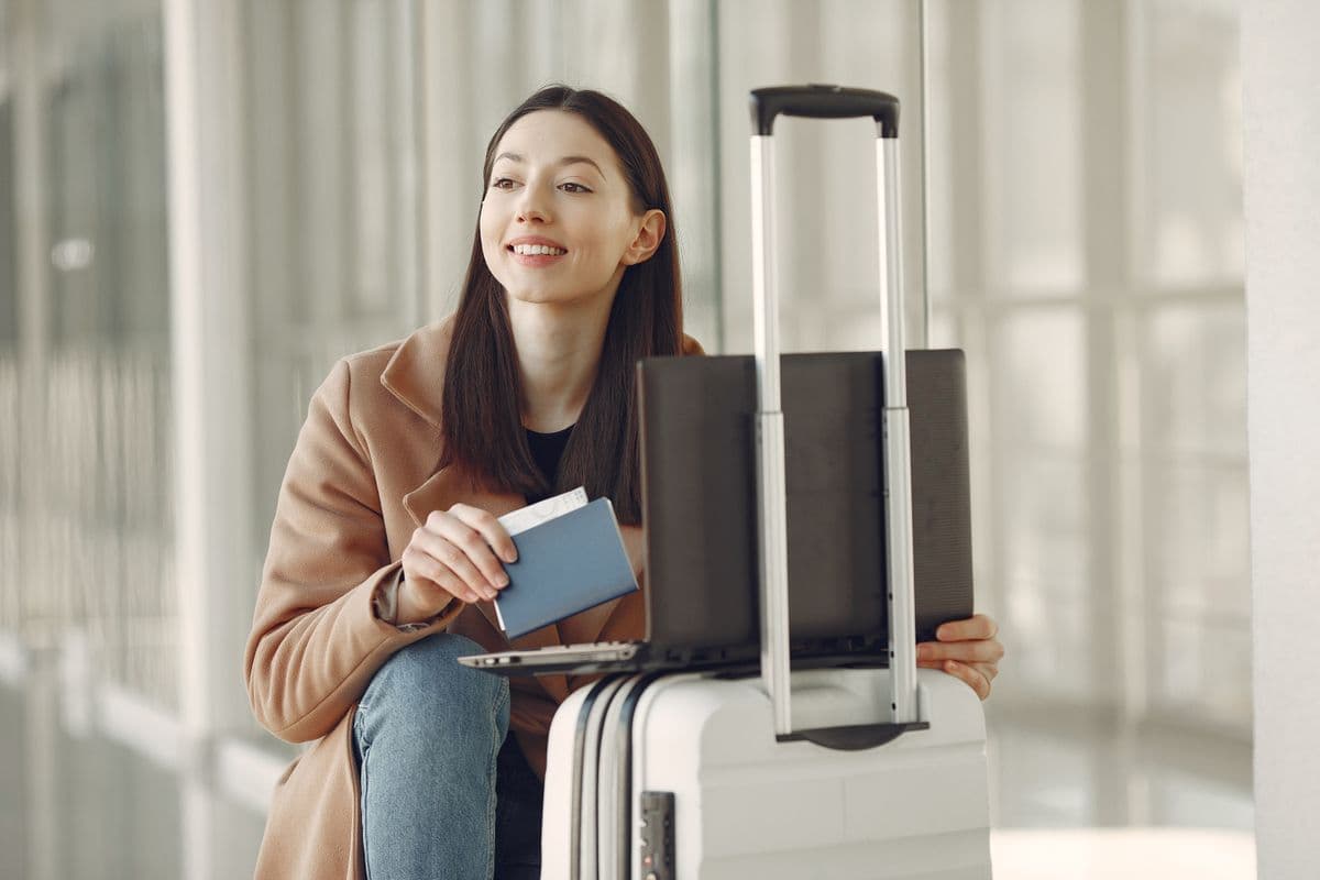 Smiling traveller holding a passport and boarding pass beside her suitcase in a bright terminal